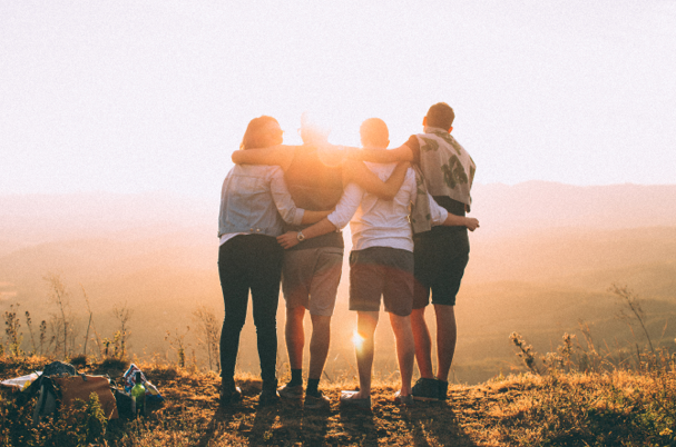 A group of friends stand at an overlook watching the sunset with their arms around each other's shoulders