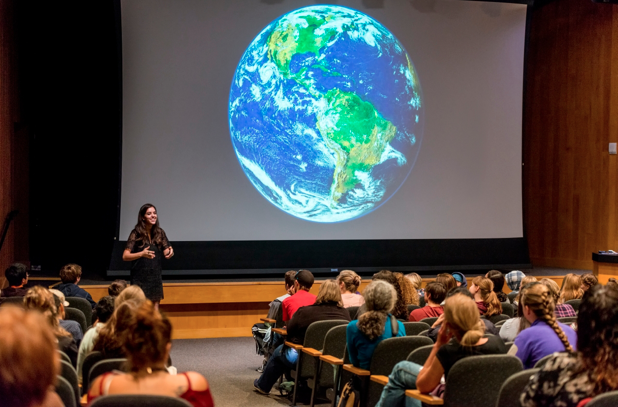 A woman addresses an audience in an auditorium with a view of Earth from space on the screen behind her