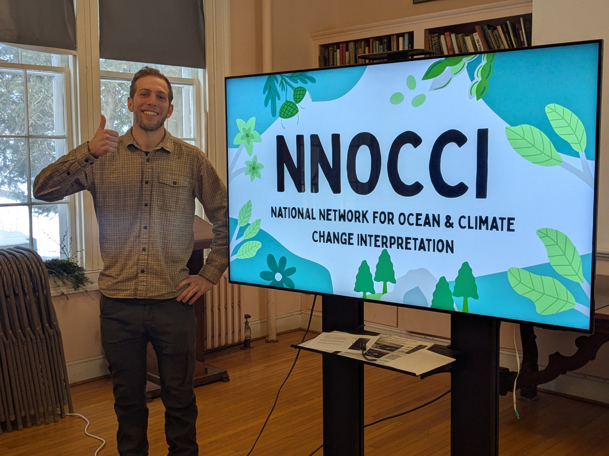 Jake poses with a thumbs up and smile beside the presentation screen that reads, "NNOCCI".
